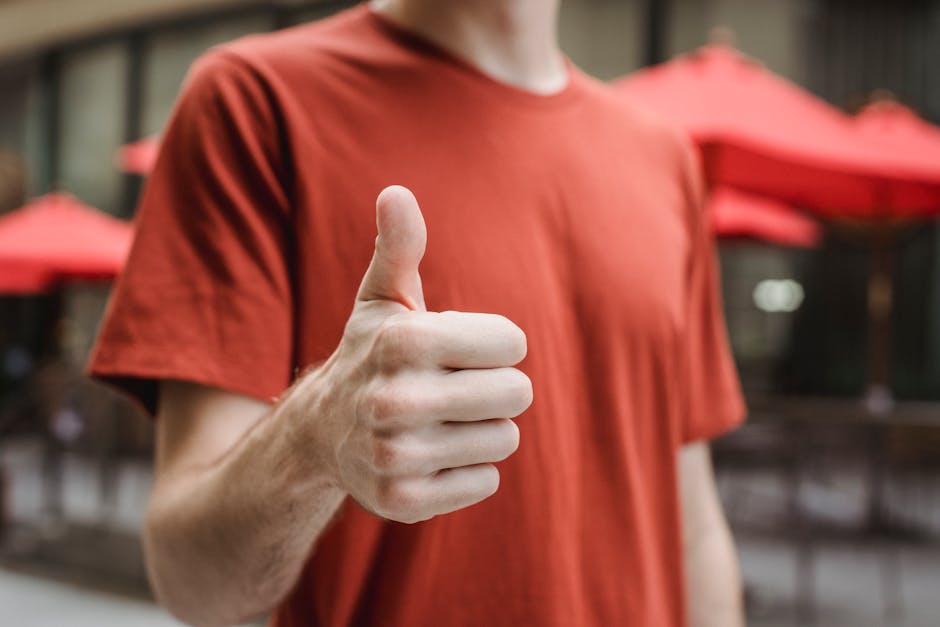 Crop anonymous young male in red t shirt showing thumb up gesture while standing on city street on sunny day